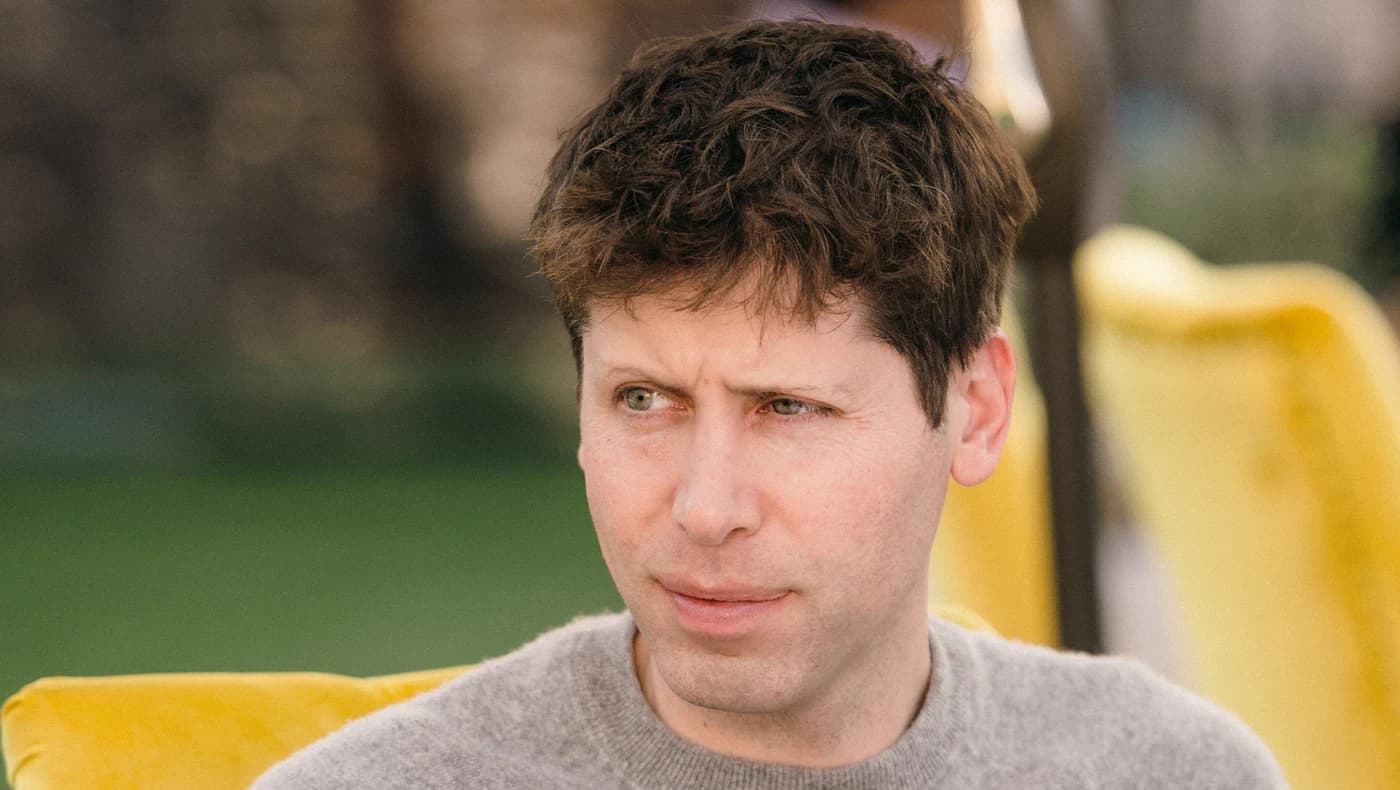 Sam Altman in a grey jumper sits at an outdoor table looking to one side